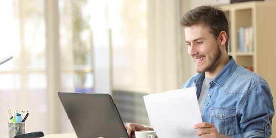 Ung glad man sitter vid sin dator hemma med papper i handen.