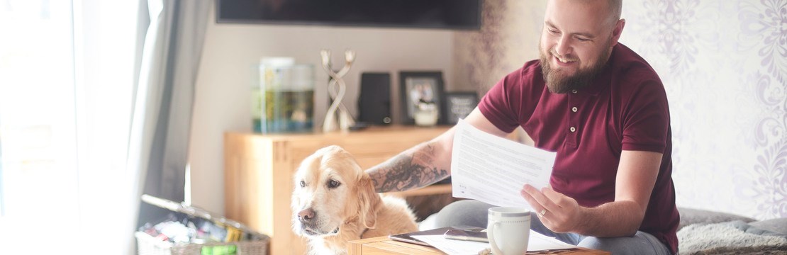 Leende man sitter hemma i soffan och håller ett papper i ena handen samtidigt som han klappar sin hund. 