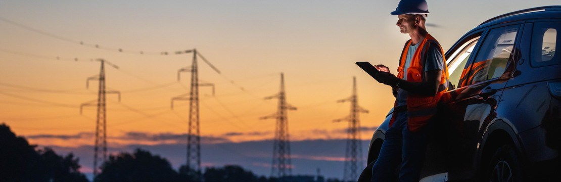En elinstallatör står lutad mot en bil med en läsplatta i handen. I bakgrunden syns kraftledningsstolpar i solnedgång.