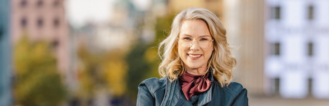 Kvinna med glasögon och rock framför en stadsmiljö.