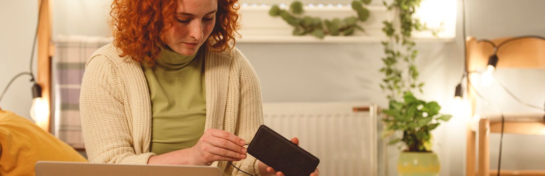 Ung kvinna  sitter i sängen i ett väl upplyst sovrum med laptop och mobiltelefon.