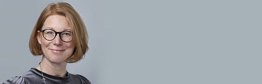 Kvinna med kort hår och glasögon framför en grå bakgrund