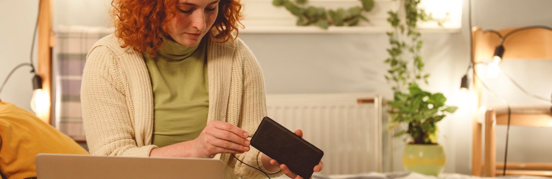 Ung kvinna  sitter i sängen i ett väl upplyst sovrum med laptop och mobiltelefon.