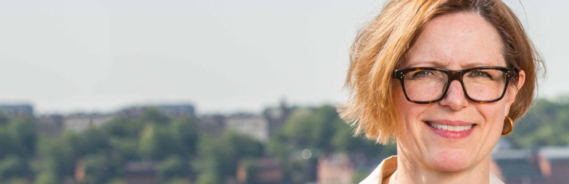 Kvinna med glasögon framför stadsmiljö med blå himmel