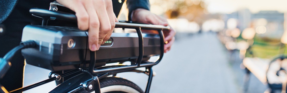 Del av elcykel samt batteri och en kostymklädd person som förbereder cykeln för avfärd i en stadsmiljö.