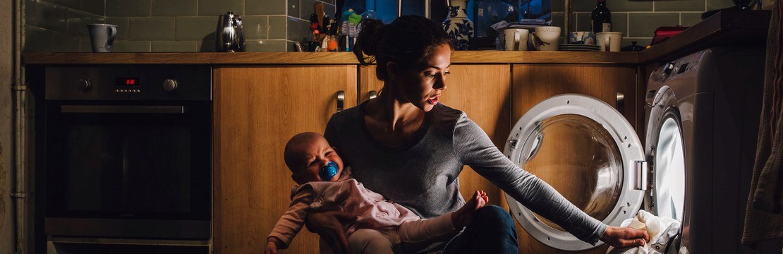 Kvinna med barn på armen sätter igång en tvättmaskin, skymning syns i fönstret.