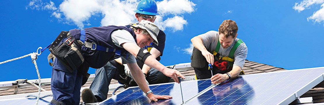 Tre män i arbetskläder och skyddslinor installerar solpaneler på tak
