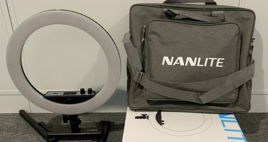Rund selfielampa i vit plast på svart fot intill en grå väska med vit text, Nanlite. Väskan står på en vit kartong.